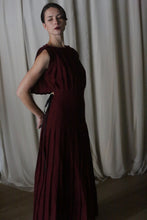 Load image into Gallery viewer, A person in a Hand Pleated Column Dress made of Japanese Wool Gauze Wine stands in profile against white curtains. Dim lighting highlights the dress&#39;s texture as they pose gracefully, one hand on their waist and the other behind.