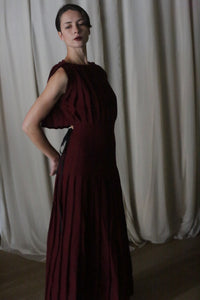 A person in a Hand Pleated Column Dress made of Japanese Wool Gauze Wine stands in profile against white curtains. Dim lighting highlights the dress's texture as they pose gracefully, one hand on their waist and the other behind.