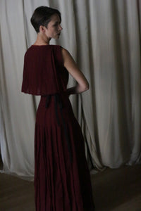 A woman, wearing the Hand Pleated Column Dress in Japanese Wool Gauze Wine, stands with her back to the camera. She has short hair and slightly turns to the side, set against beige curtains.