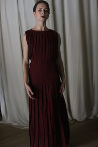 In a dimly lit room with beige curtains, a person with short dark hair stands in a Hand Pleated Column Dress in Japanese Wool Gauze Wine. Their arms at their sides and neutral expression highlight the elegance of the dark red, pleated dress.