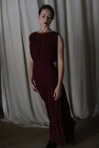 In a dimly lit room, a person elegantly wears the Hand Pleated Column Dress in rich wine-colored Japanese wool gauze. Soft, flowing curtains in the background enhance the scene's refined ambience.