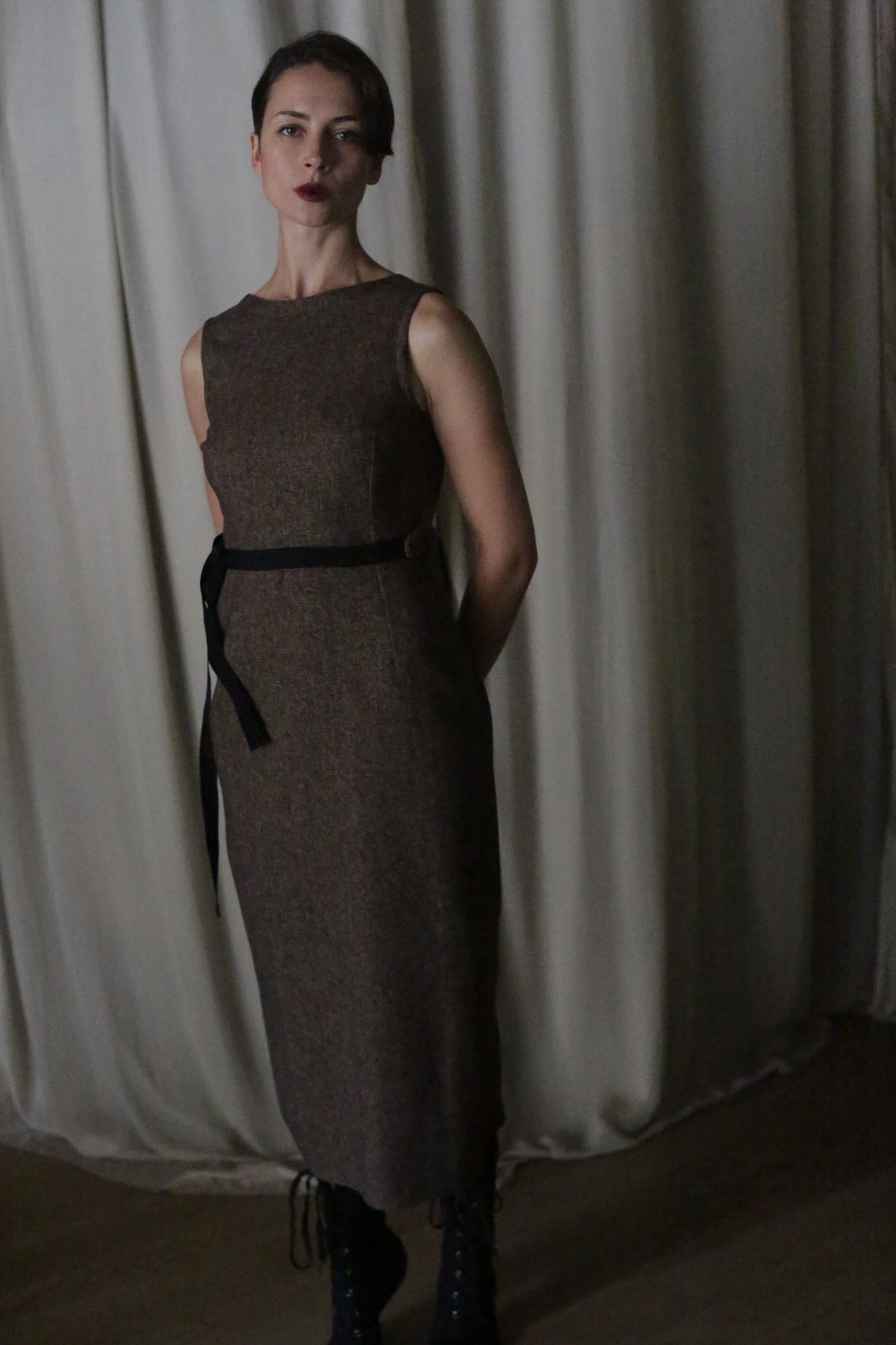 In dim lighting, a woman with dark hair and red lipstick stands in front of a white curtain. She wears the sleeveless "Drape Dress" made from brown cotton twill, paired with a black belt and matching boots. Her arms are positioned behind her back as subtle shadows are cast around her.