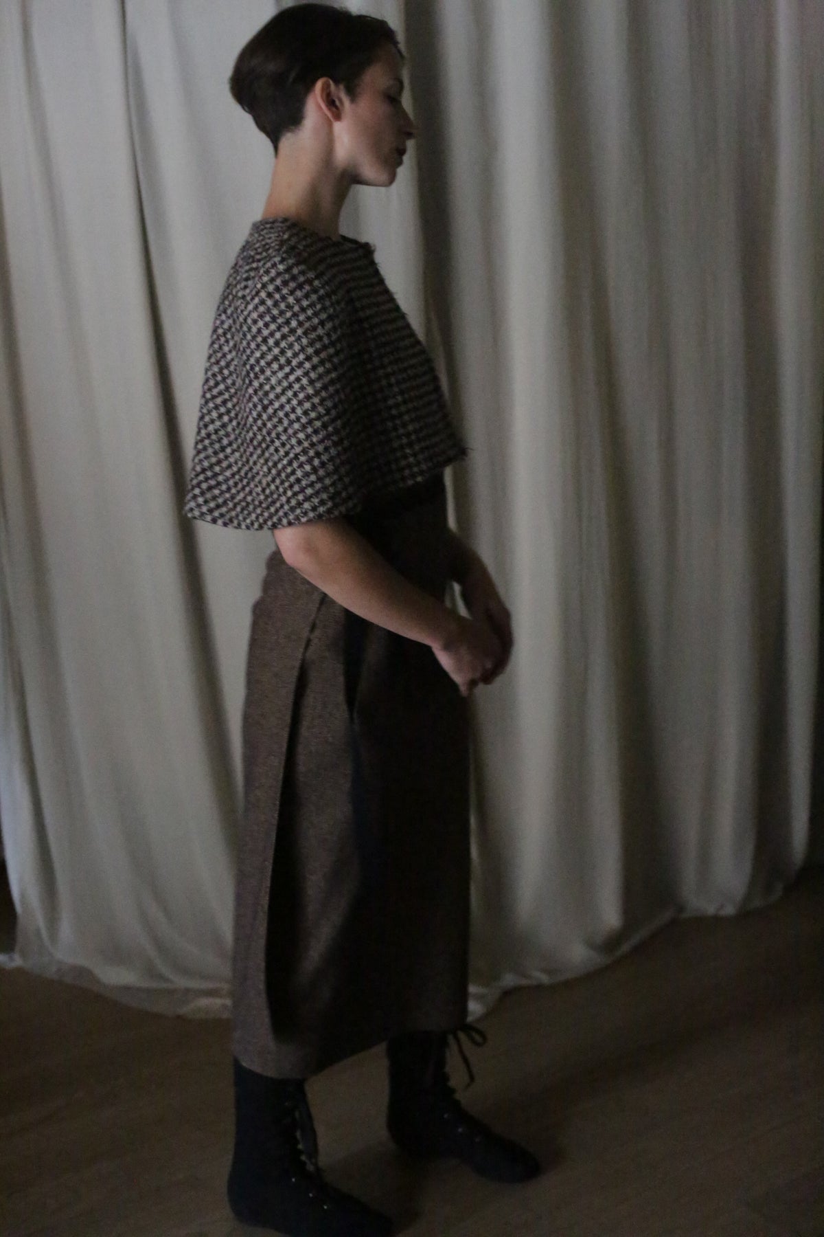 A person with short hair stands in profile against a curtain, wearing a patterned top, long brown skirt, black lace-up boots, and the Alpaca Capelet, with hands gently clasped in front.