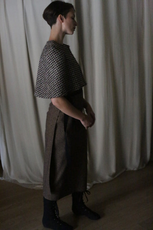 A person with short hair stands in profile against a curtain, wearing a patterned top, long brown skirt, black lace-up boots, and the Alpaca Capelet, with hands gently clasped in front.
