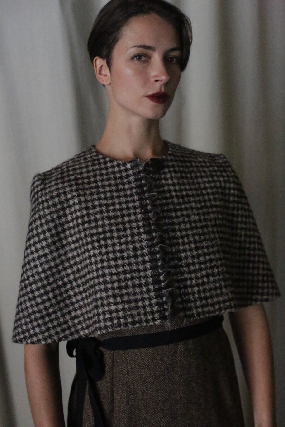 A woman with short dark hair stands against a soft, light backdrop wearing the handmade in San Francisco Alpaca Capelet with a ruffled front over a belted brown dress, gazing confidently at the camera.