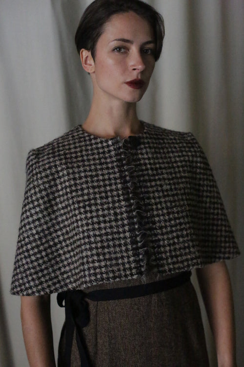 A woman with short dark hair stands against a soft, light backdrop wearing the handmade in San Francisco Alpaca Capelet with a ruffled front over a belted brown dress, gazing confidently at the camera.
