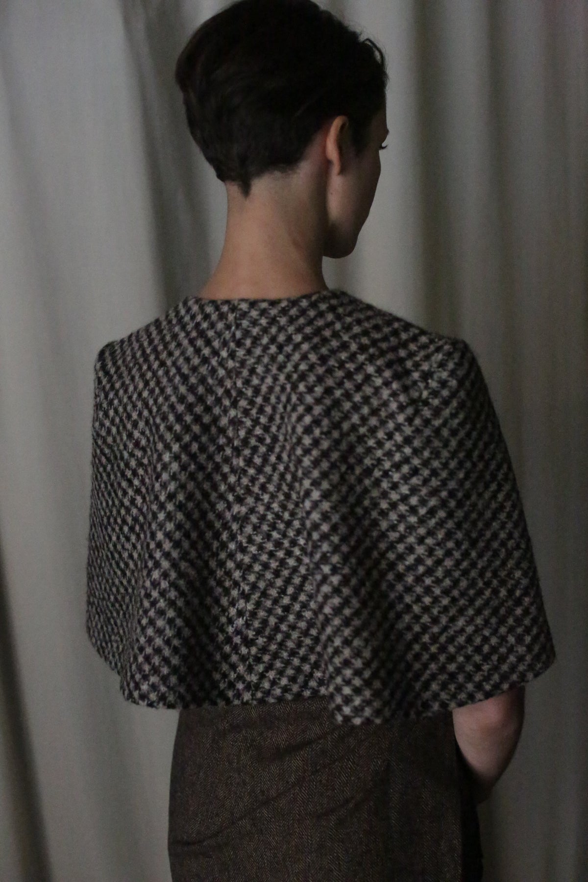 A person with short dark hair faces away, wearing the Alpaca Capelet—handmade in San Francisco from Italian alpaca bouclé—against a soft, neutral backdrop.