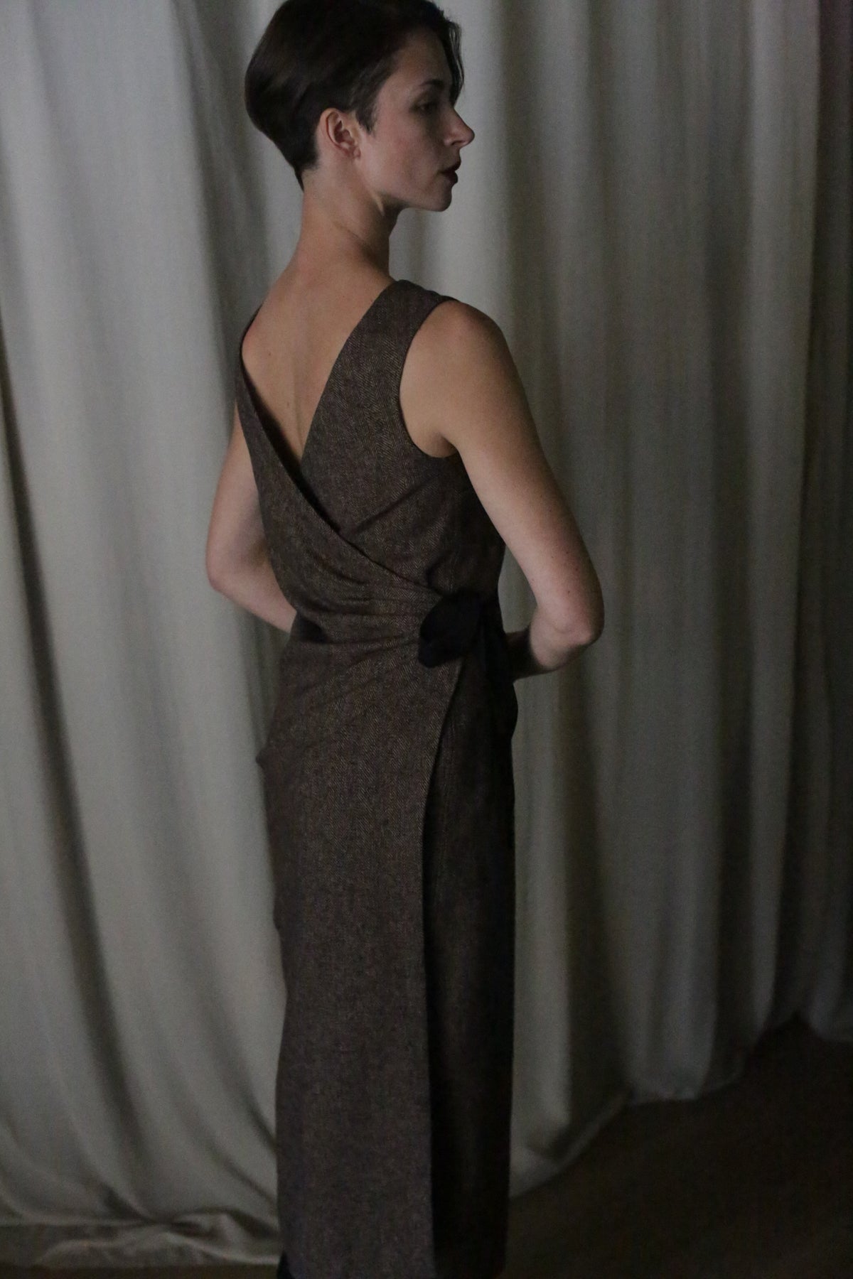 In a dimly lit room, a woman with short dark hair wears the elegant sleeveless Drape Dress, featuring a deep V-neck and backless design. Made from luxurious cotton twill, this reversible piece drapes gracefully against light-colored curtains in the background.