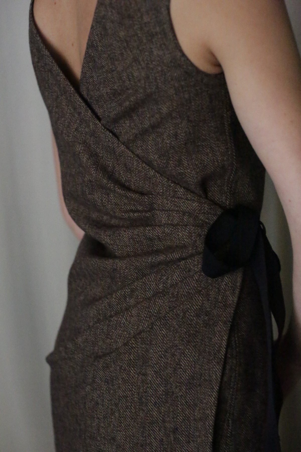 A close-up of someone wearing the Drape Dress shows its brown textured fabric and a black belt at the waist. The sleeveless design highlights its elegant, reversible draping from the back, emphasizing its exquisite details.