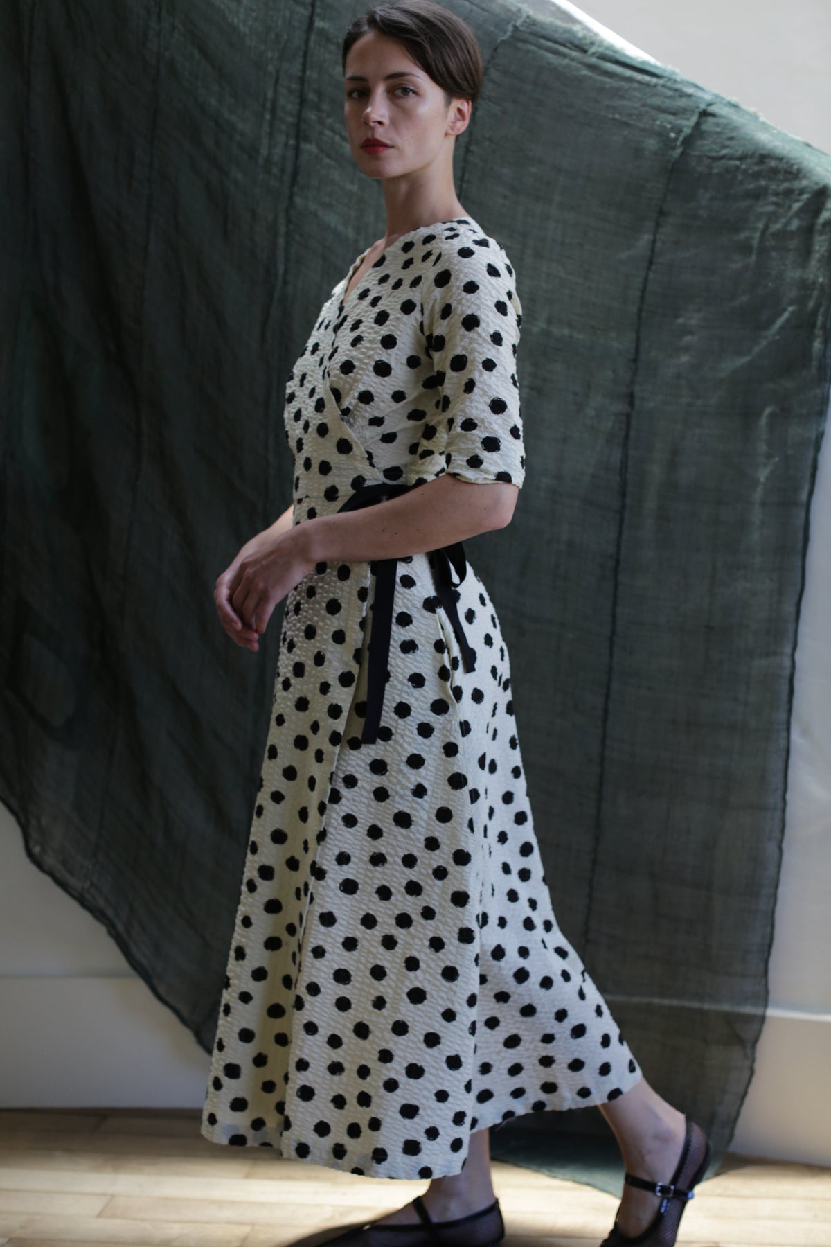 A woman wears the Fitted Wrap Dress | Polkadot Seersucker Ivory, featuring a black-on-ivory polkadot pattern. Indoors on a wooden floor with a dark fabric backdrop, she looks over her shoulder, her hair simply pulled back.