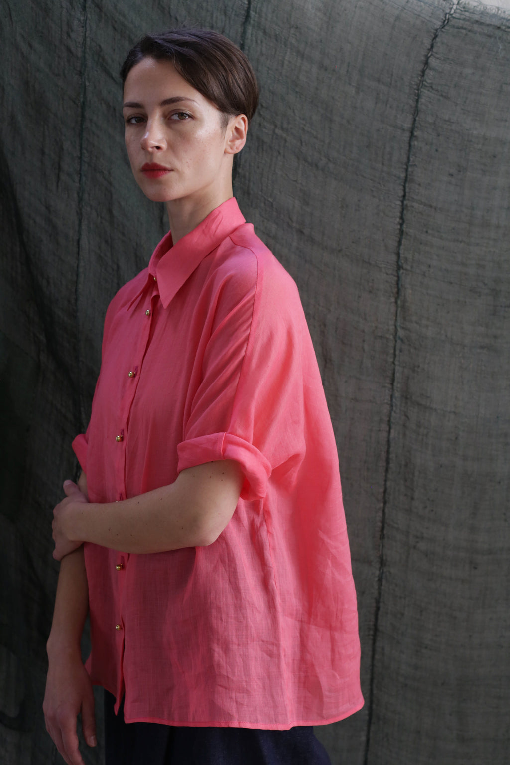 A person with short dark hair, wearing the Work Shirt | Ramie Sakura—a loose-fitting pink shirt with rolled sleeves and brass buttons—stands before a textured gray-green backdrop, looking directly at the camera with a neutral expression.