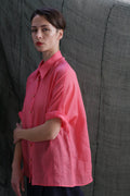 A person with short dark hair, wearing the Work Shirt | Ramie Sakura—a loose-fitting pink shirt with rolled sleeves and brass buttons—stands before a textured gray-green backdrop, looking directly at the camera with a neutral expression.