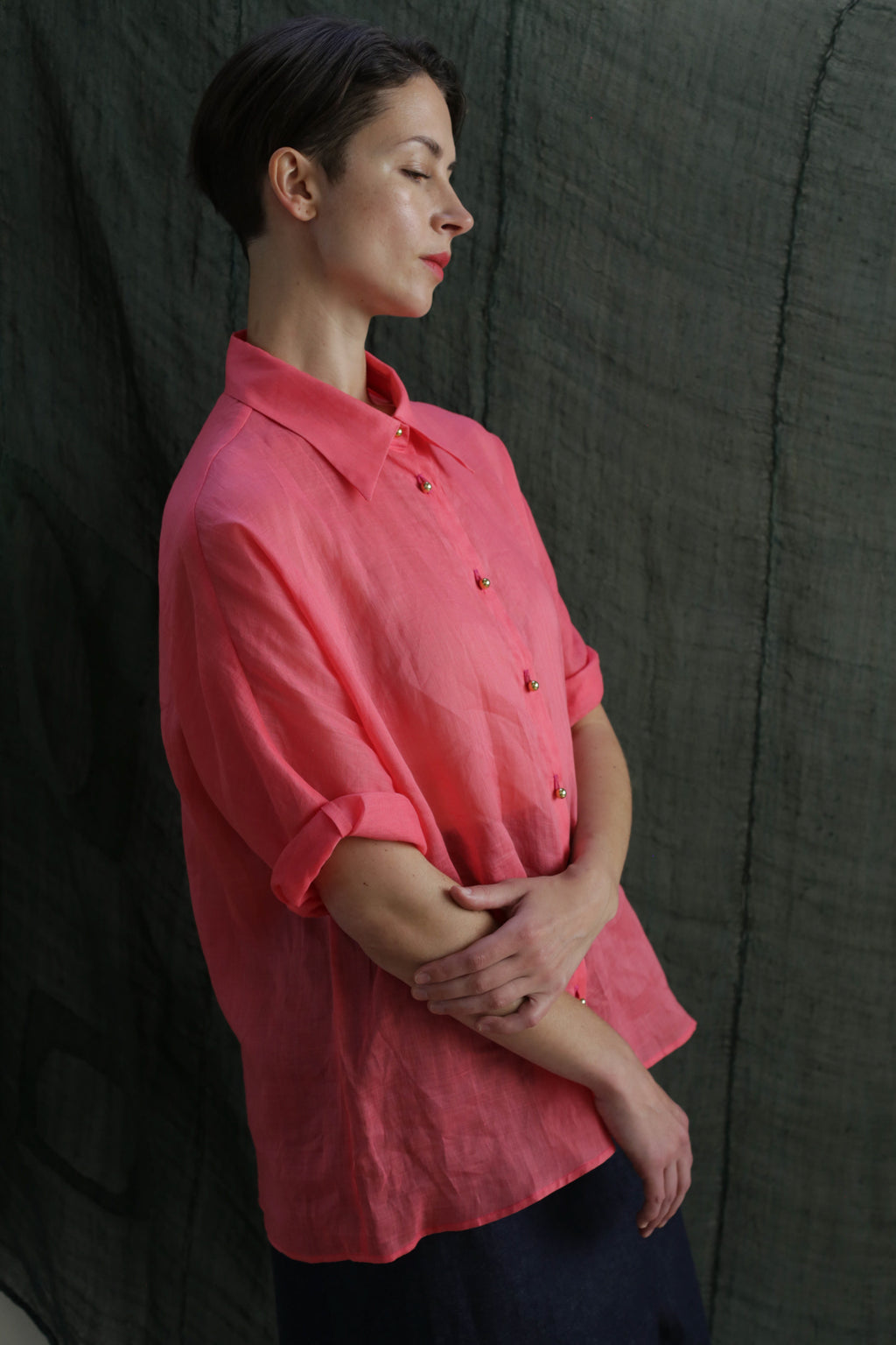 A woman with short brown hair stands in profile, arms folded and eyes closed, wearing the Work Shirt | Ramie Sakura—a loose, coral pink classic ramie shirt with brass buttons—against a dark green fabric backdrop.