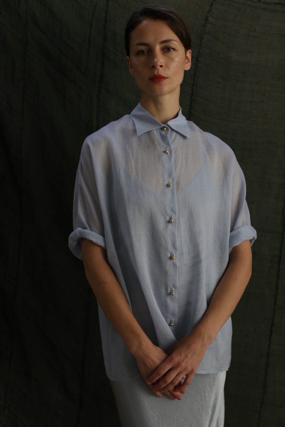 A woman stands before a dark green fabric backdrop, hands folded and expression neutral, wearing the Work Shirt | Ramie Mist—a sheer, light blue ramie button-up.