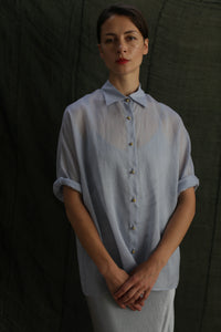 A woman stands before a dark green fabric backdrop, hands folded and expression neutral, wearing the Work Shirt | Ramie Mist—a sheer, light blue ramie button-up.