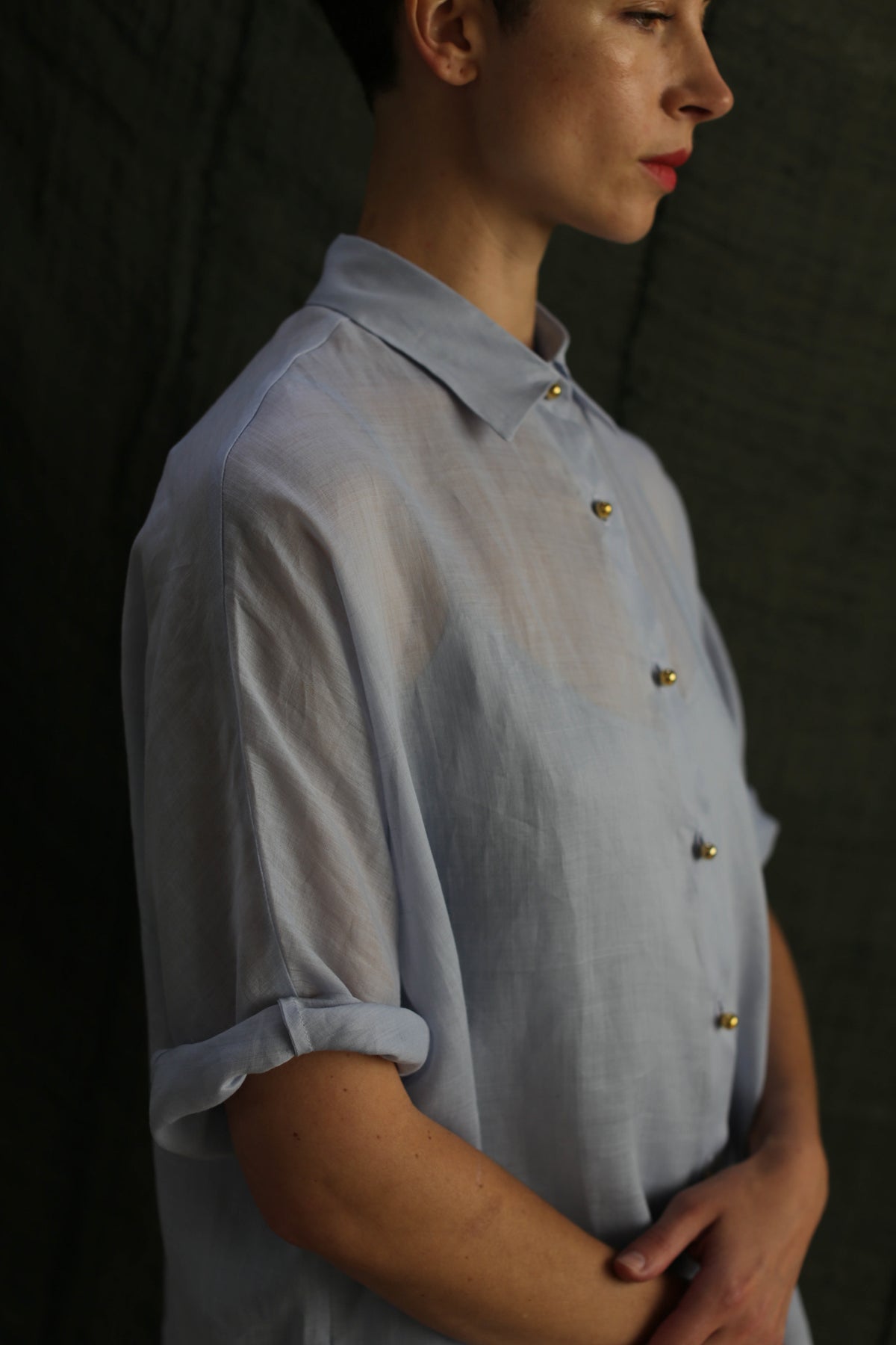 A person in a light blue Work Shirt | Ramie Mist with rolled sleeves and brass buttons stands against a dark background. The photo is cropped above the nose, leaving the person's eyes out of view.