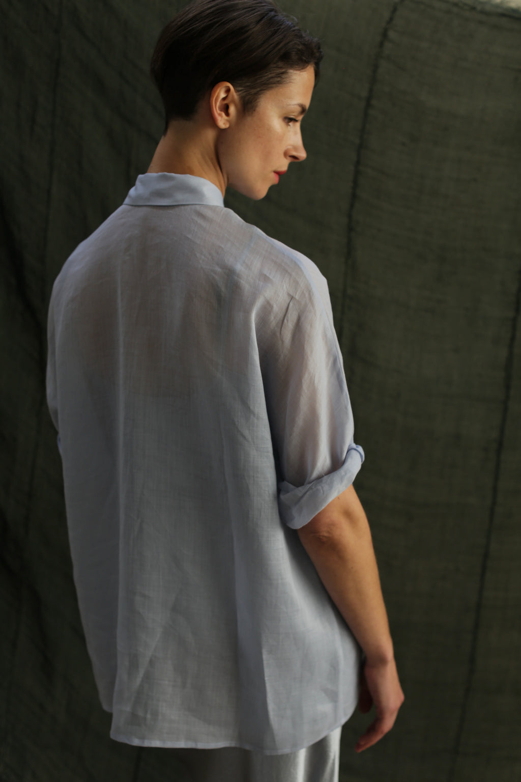 A person with short brown hair wears the Work Shirt | Ramie Mist, a loose, light-hued top with rolled-up sleeves, standing against a dark green textured backdrop and looking to the side.