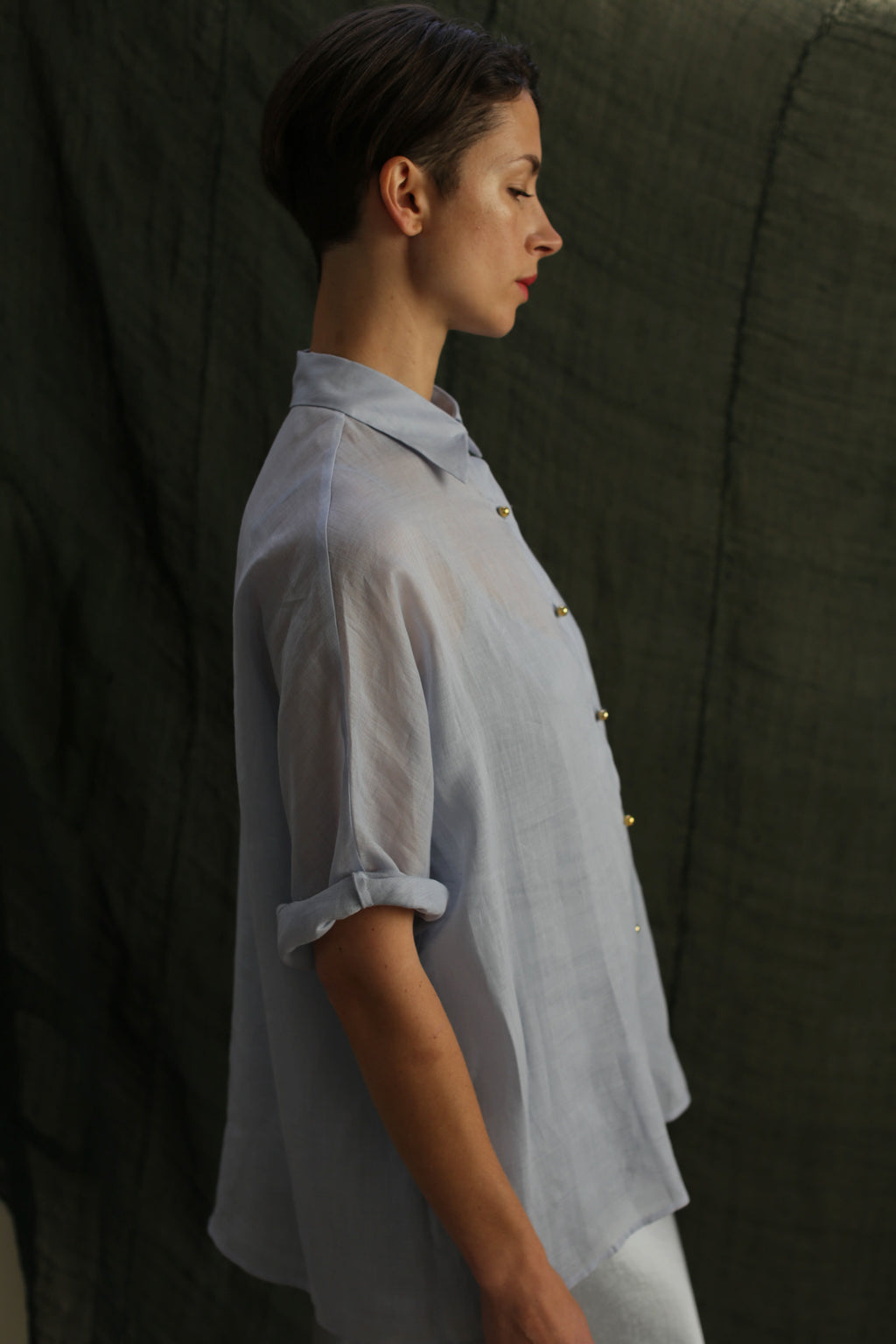 A person with short hair stands in profile against a dark fabric background, wearing the Work Shirt | Ramie Mist with rolled sleeves and subtle brass buttons.