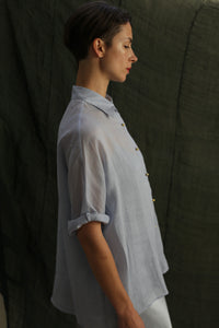 A person with short hair stands in profile against a dark fabric background, wearing the Work Shirt | Ramie Mist with rolled sleeves and subtle brass buttons.