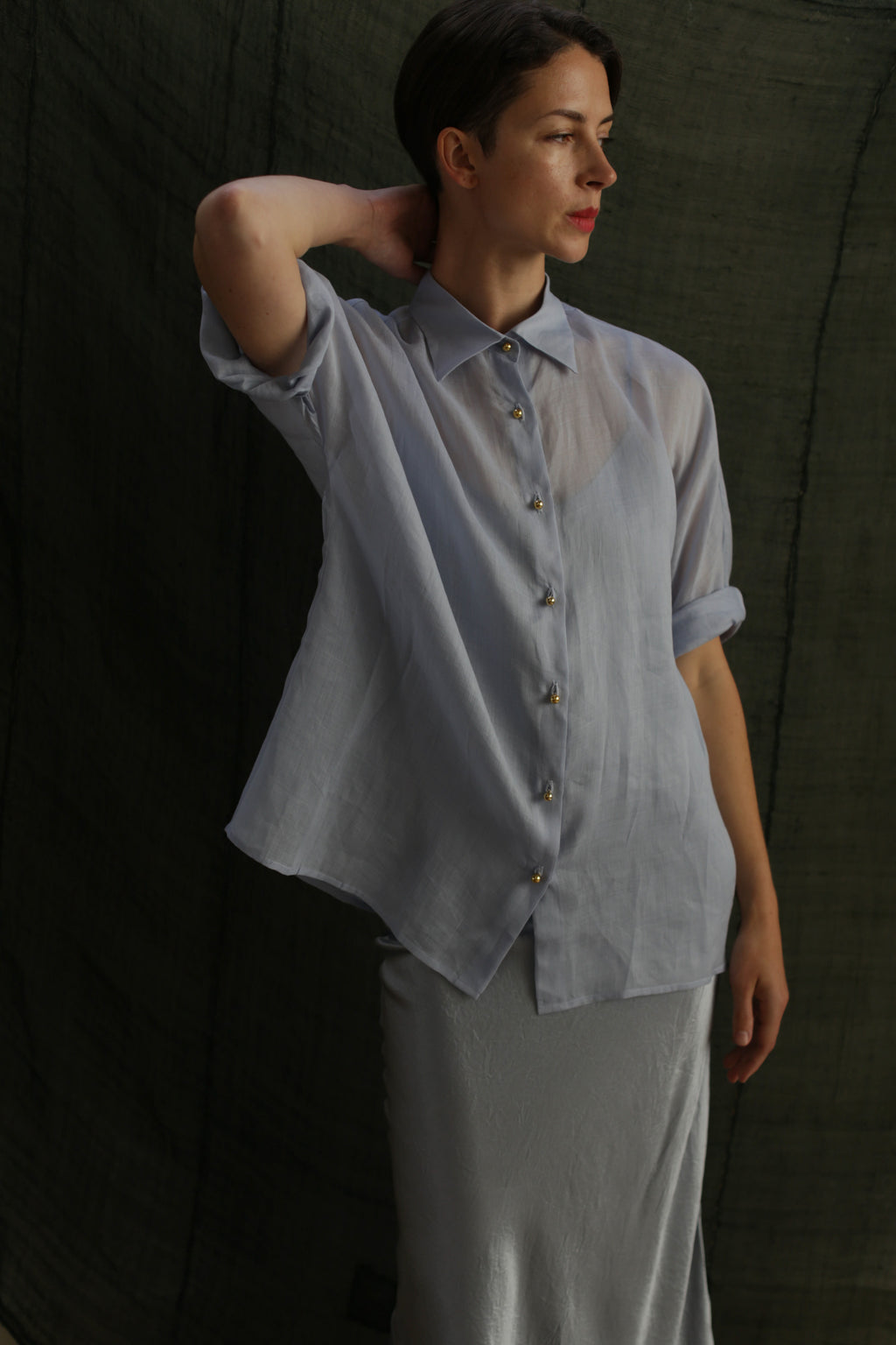 A woman wears the Work Shirt | Ramie Mist, a sheer light blue top with brass buttons, paired with a light grey skirt. She stands against a dark textured background, one arm bent and resting on her head as she looks aside.