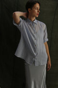 A woman wears the Work Shirt | Ramie Mist, a sheer light blue top with brass buttons, paired with a light grey skirt. She stands against a dark textured background, one arm bent and resting on her head as she looks aside.