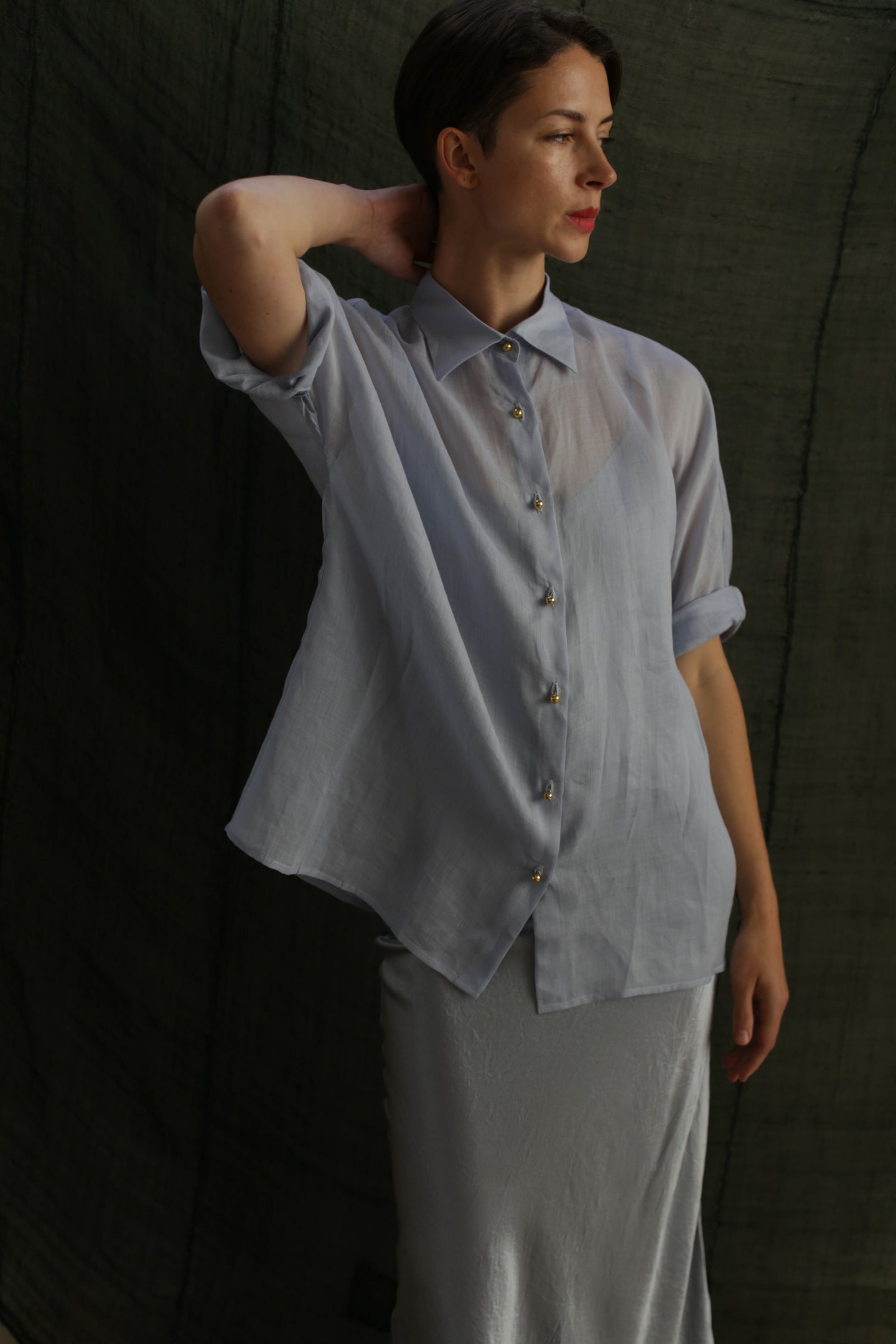 A woman wears the Work Shirt | Ramie Mist, a sheer light blue top with brass buttons, paired with a light grey skirt. She stands against a dark textured background, one arm bent and resting on her head as she looks aside.