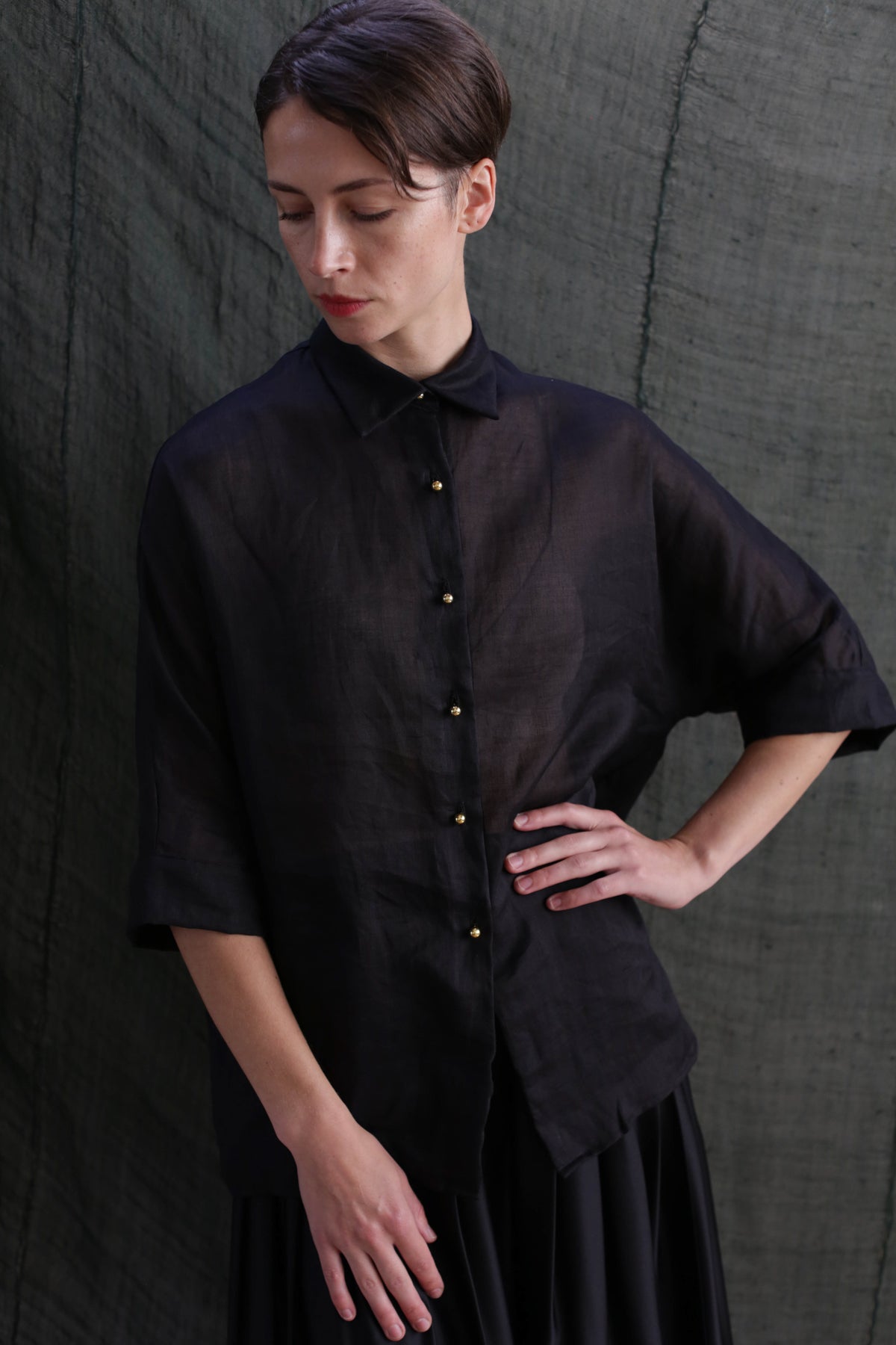 A woman with short brown hair stands against a dark textured background, wearing the Work Shirt | Ramie Black with solid brass buttons and elbow-length sleeves, paired with a black skirt, looking downward with one hand on her hip.