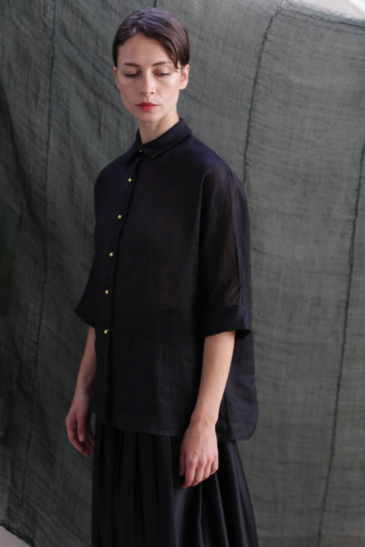 A woman stands before a textured green backdrop, wearing the Work Shirt | Ramie Black with solid brass buttons and a matching black skirt. She gazes downward, her expression neutral.