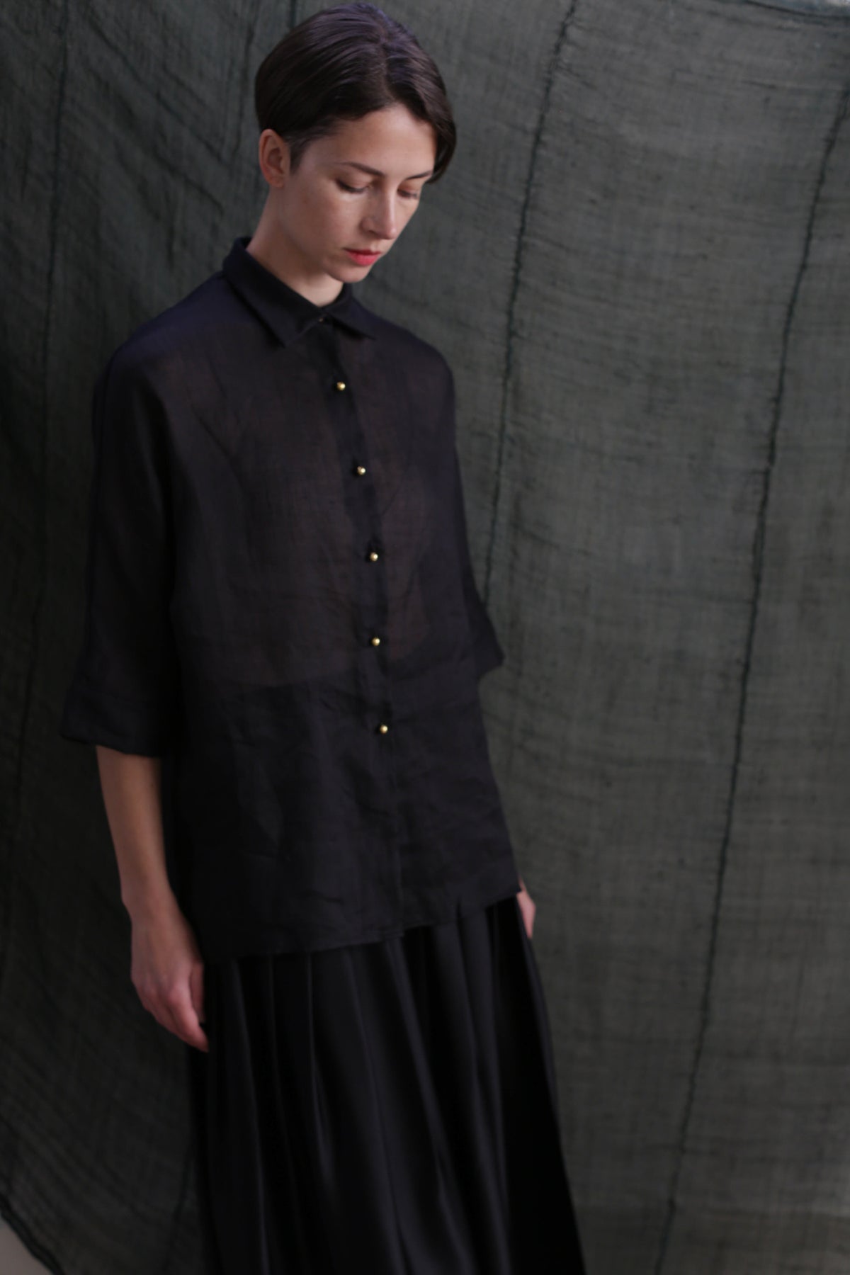 A person with short hair wears the Work Shirt | Ramie Black, featuring solid brass buttons, and a black skirt, standing against a textured dark fabric backdrop and looking downward with a calm expression.