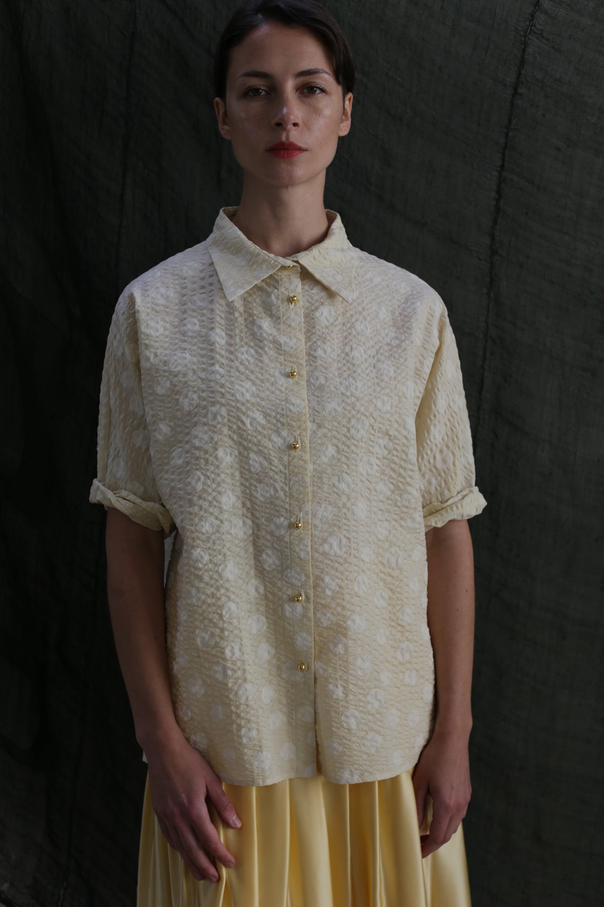 A woman stands against a dark textured background, wearing the Work Shirt | Polkadot Seersucker Butter—a loose, short-sleeved Japanese cotton button-up with brass buttons—paired with a light yellow skirt. She looks at the camera with a neutral expression.
