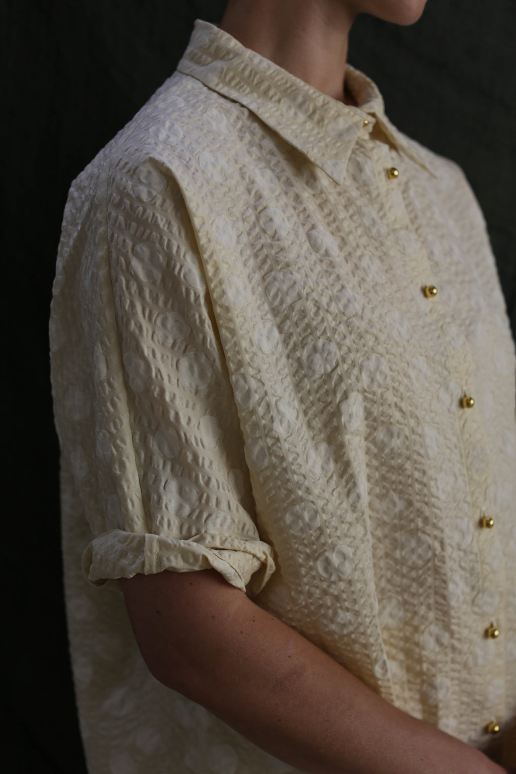A person wears the Work Shirt | Polkadot Seersucker Butter, a textured cream button-up with brass buttons and rolled sleeves. Only their torso and lower face are visible against a dark background.