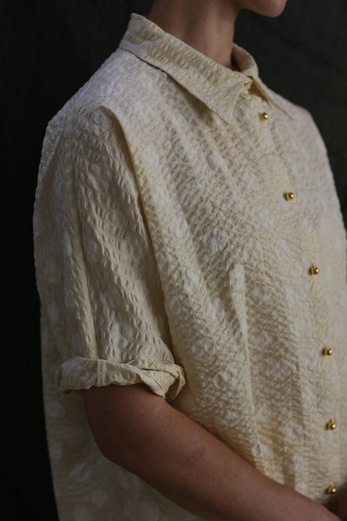 A person wears the Work Shirt | Polkadot Seersucker Butter, a textured cream button-up with brass buttons and rolled sleeves. Only their torso and lower face are visible against a dark background.