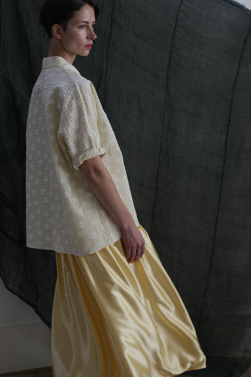 A person in the Work Shirt | Polkadot Seersucker Butter stands sideways against a dark, sheer backdrop, gazing thoughtfully into the distance.