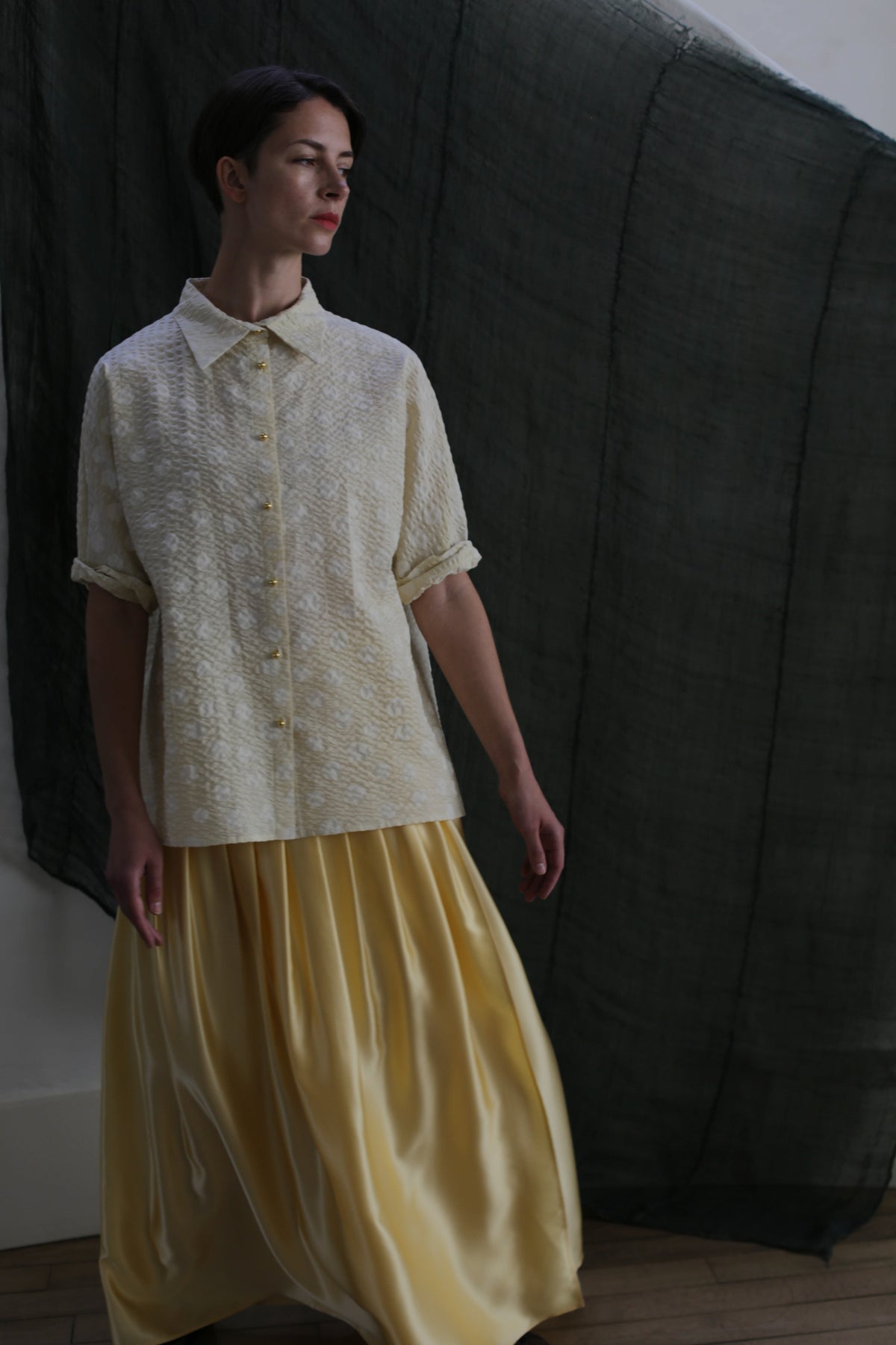 A person wearing a cream textured short-sleeve blouse and the Hand Pleated Column Wrap Skirt in Silk Charmeuse Butter stands against a dark fabric backdrop, gazing slightly to the side.