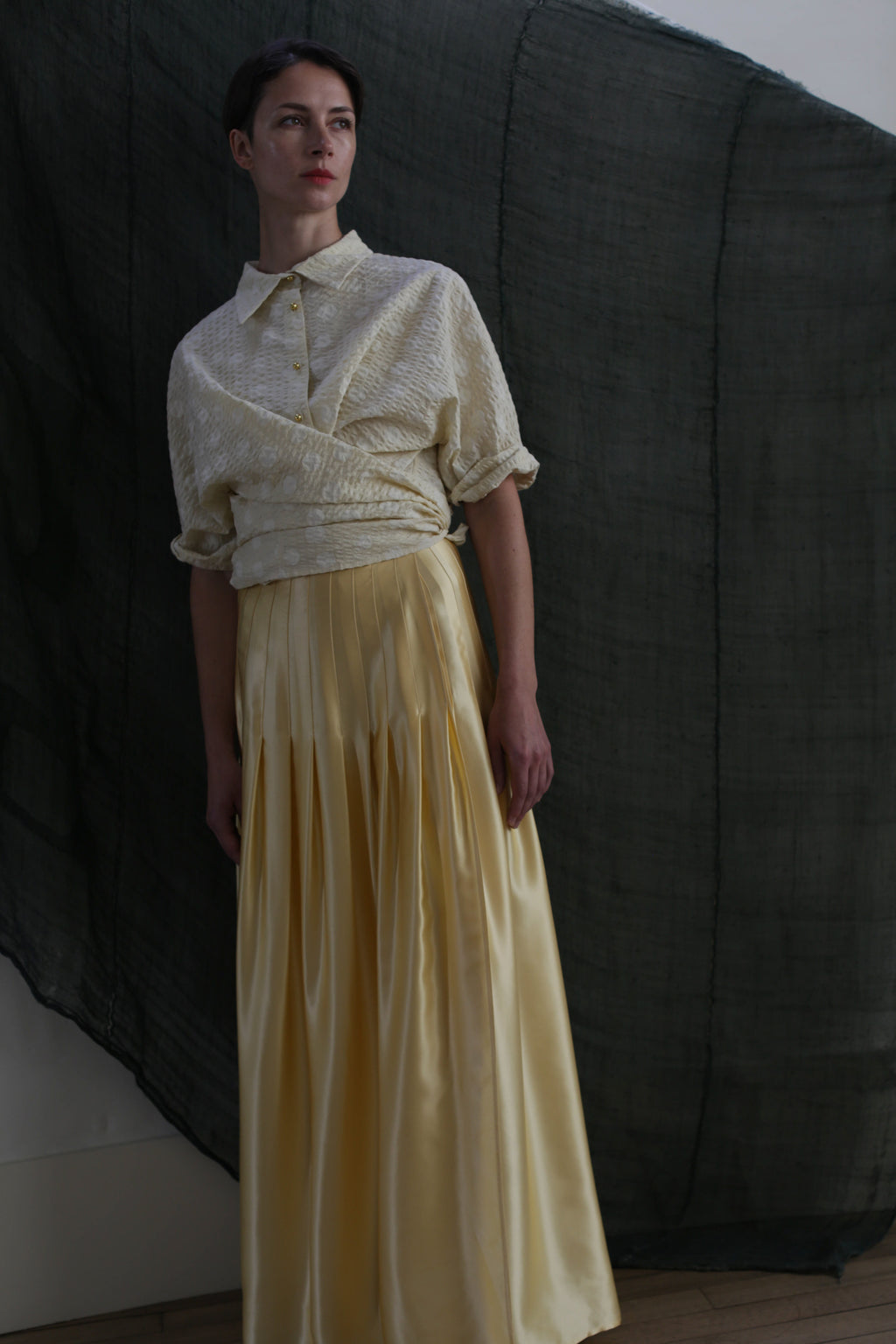 A person stands against a dark fabric backdrop, wearing the Work Shirt in Polkadot Seersucker Butter with rolled sleeves and a long, flowing yellow satin skirt. Soft, subdued lighting creates a moody atmosphere.