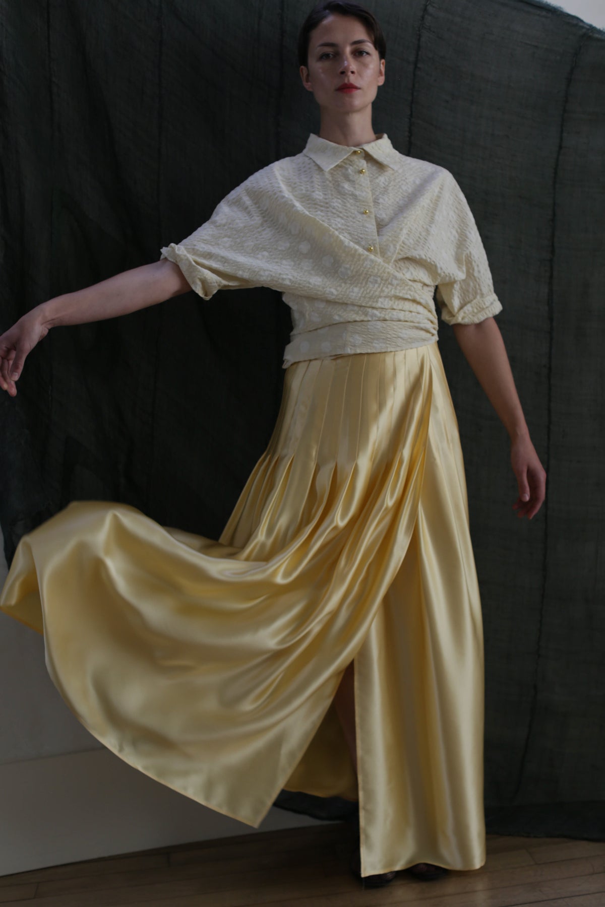 Against a dark fabric backdrop, a person poses gracefully with one arm extended, wearing a cream textured blouse and the Hand Pleated Column Wrap Skirt | Silk Charmeuse Butter, the skirt flowing and billowing elegantly.