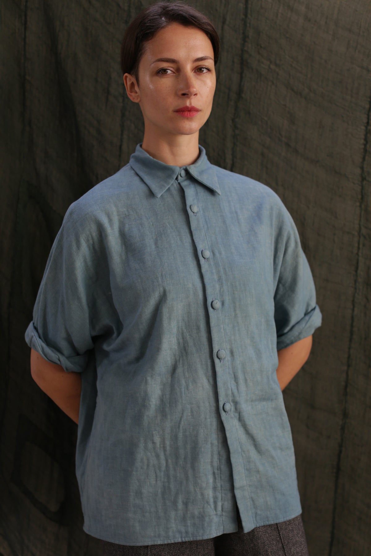 A woman with short brown hair stands before a dark background, wearing the Work Shirt | Triple Gauze Ai Dye—a loose indigo button-up featuring rolled sleeves and hand-sewn buttons, her hands tucked behind her back.