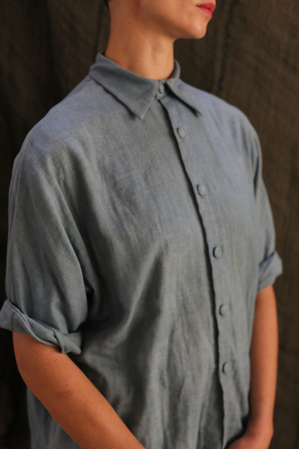 A person in the Work Shirt | Triple Gauze Ai Dye—an indigo-dyed, loose-fit button-up with hand-sewn buttons and rolled sleeves—stands against a dark, textured background, their face partly out of frame.