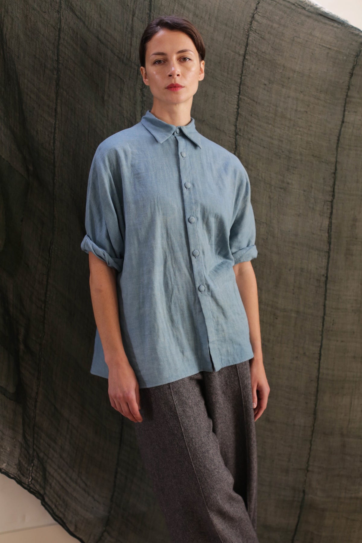 A woman stands before a textured dark green backdrop, wearing the Work Shirt | Triple Gauze Ai Dye in light blue with rolled sleeves and loose gray pants, gazing calmly at the camera.