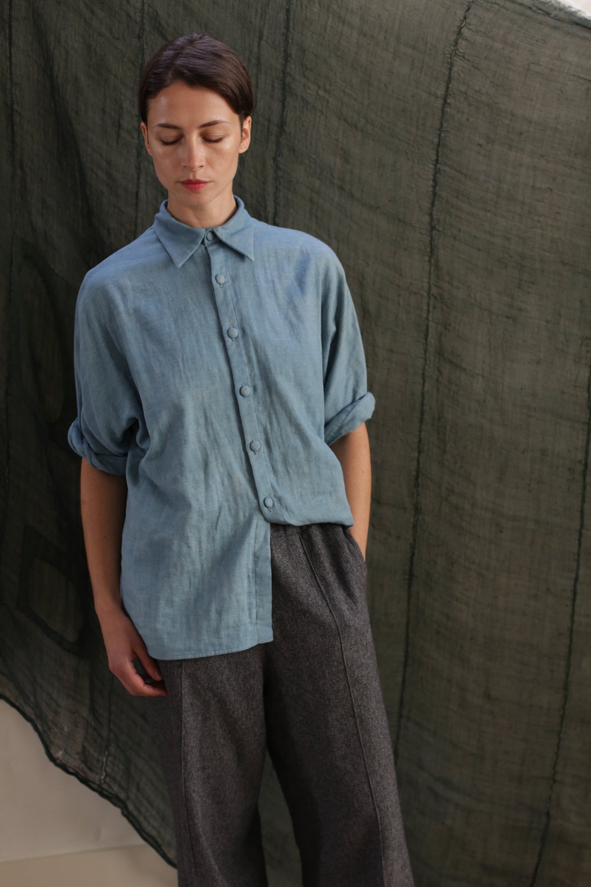 A person stands with one hand in their pocket, wearing the Work Shirt | Triple Gauze Ai Dye—a loose indigo-dyed cotton shirt with rolled sleeves—and gray pants, in front of a dark green fabric backdrop.