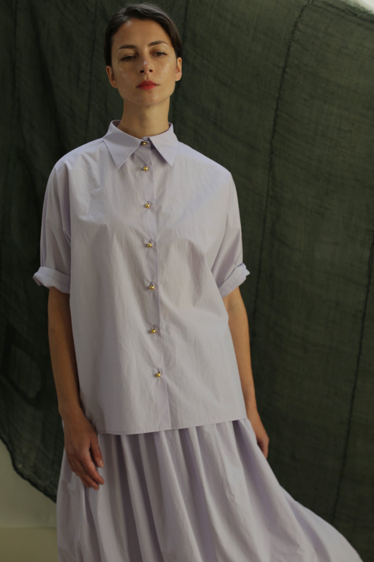 A person stands before a dark green fabric backdrop wearing the Work Shirt | Typewriter Cotton Amethyst, a loose, light lavender Japanese cotton button-up with rolled sleeves, paired with a matching pleated skirt.