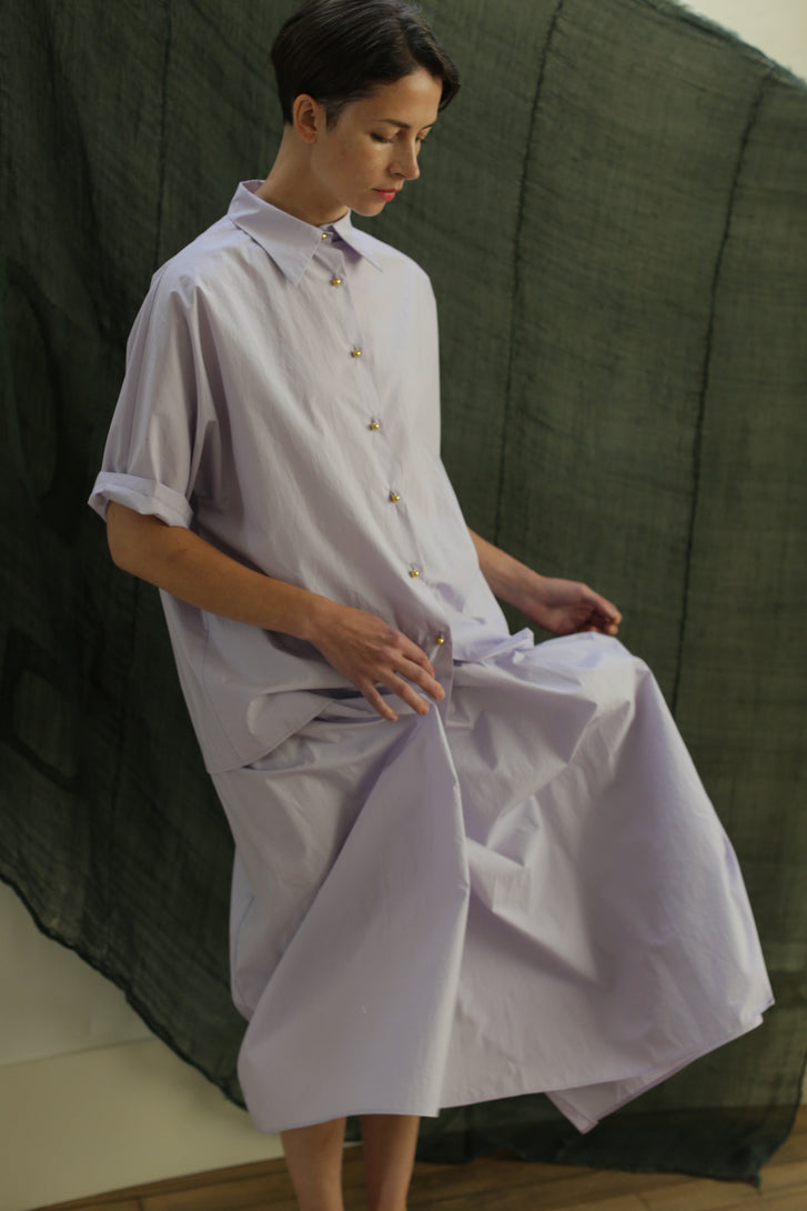 A person with short hair and a calm expression sits with one leg bent, holding the fabric of the Work Shirt | Typewriter Cotton Amethyst. A dark green draped cloth serves as the background.
