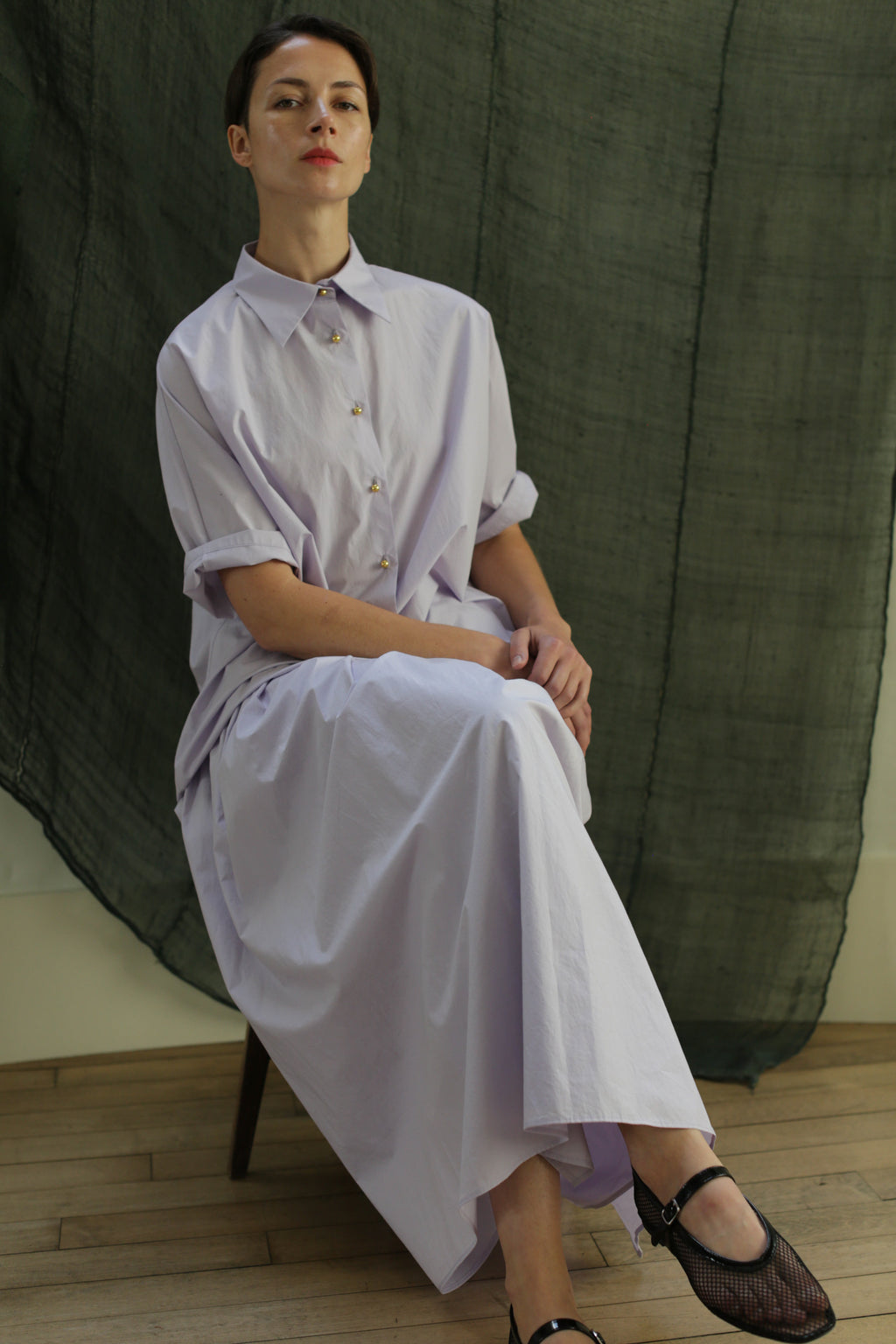 A person sits cross-legged on a chair, wearing the Work Shirt | Typewriter Cotton Amethyst—a loose, light lavender button-down made from Japanese Typewriter Cotton with brass buttons. A dark green textured fabric is draped in the background.