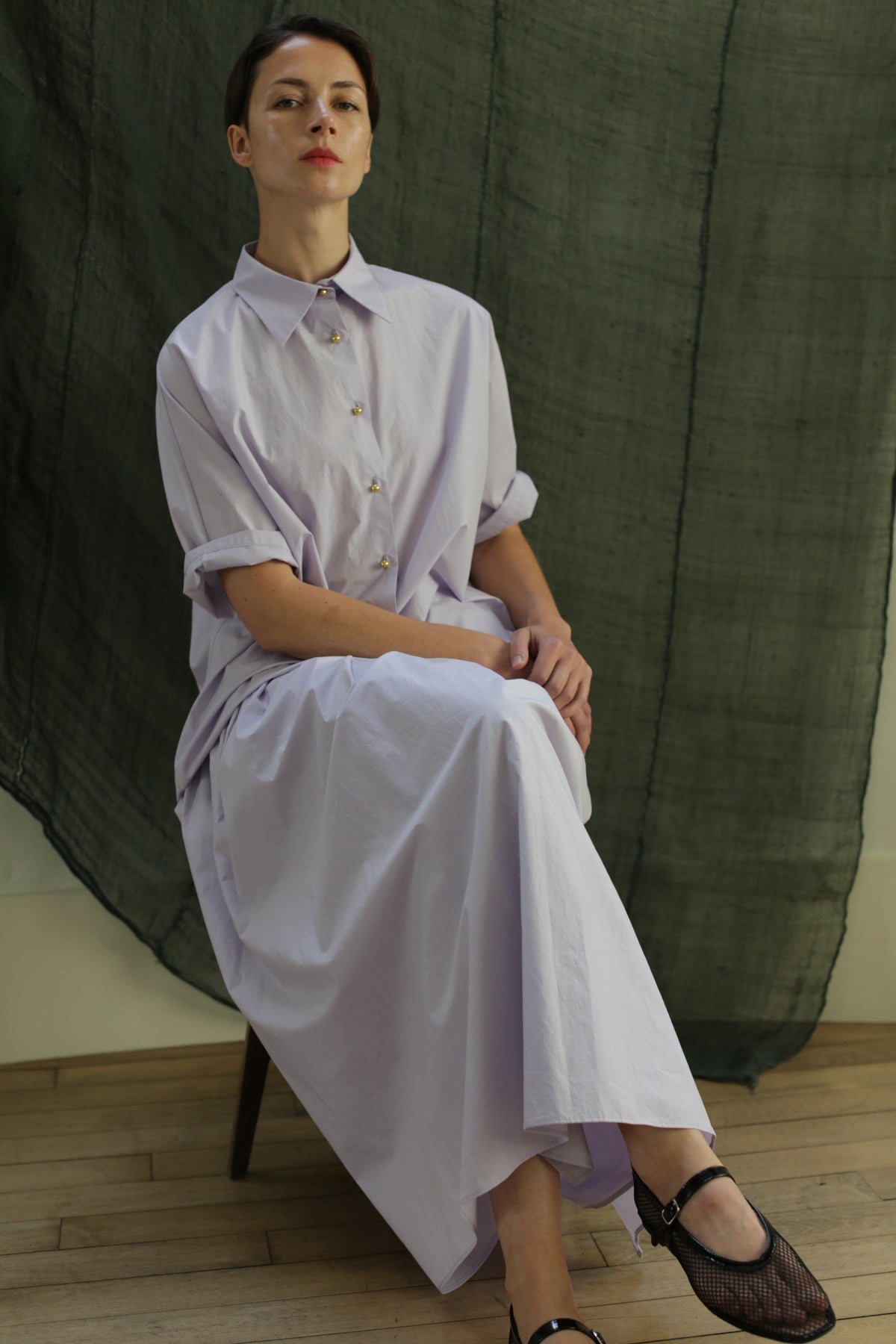 A person sits cross-legged on a chair, wearing the Work Shirt | Typewriter Cotton Amethyst—a loose, light lavender button-down made from Japanese Typewriter Cotton with brass buttons. A dark green textured fabric is draped in the background.