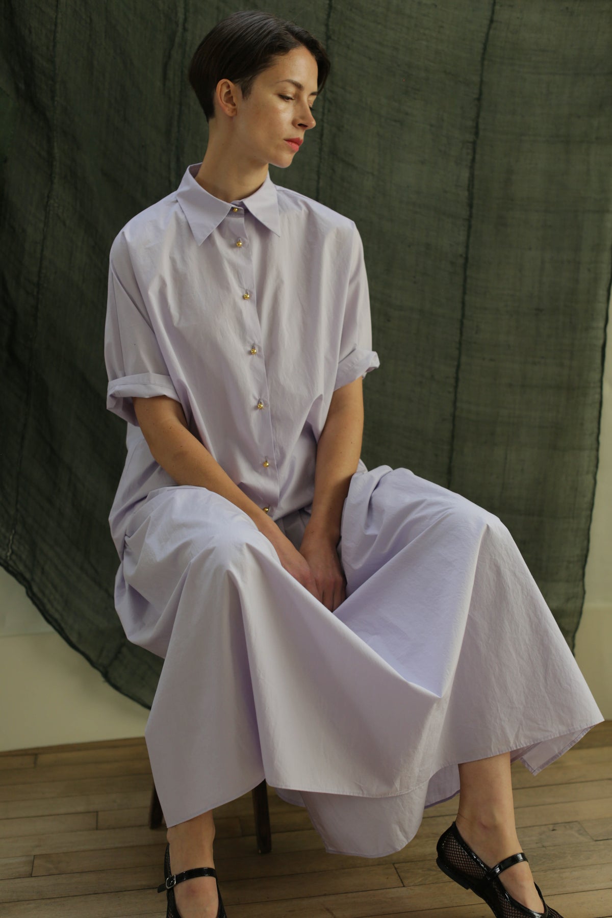 A woman in the Work Shirt | Typewriter Cotton Amethyst sits on a stool, hands in her lap, gazing to the side against a dark green textured fabric backdrop.