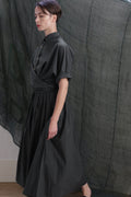 A person with short brown hair models the black Typewriter Wrap Skirt, featuring a pleated design, rolled sleeves, and a dark collar, while walking indoors beside a sheer, dark fabric backdrop.