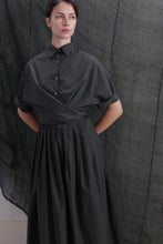 Load image into Gallery viewer, A woman stands with her hands behind her back, wearing the Work Shirt in Typewriter Cotton Black, featuring a dark collar, short sleeves, brass buttons, and a wrapped waist. The background shows draped dark green fabric.
