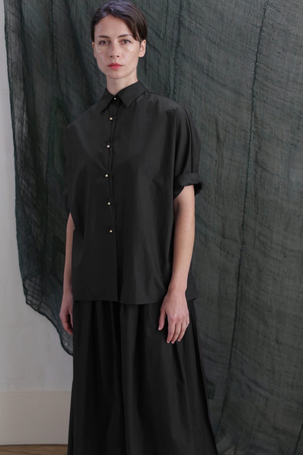 A person with short dark hair wears the Work Shirt | Typewriter Cotton Black and a black skirt with brass buttons, standing before a textured dark green fabric backdrop.