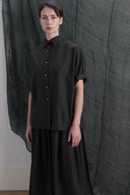 Load image into Gallery viewer, A person with short dark hair wears the Work Shirt | Typewriter Cotton Black and a black skirt with brass buttons, standing before a textured dark green fabric backdrop.