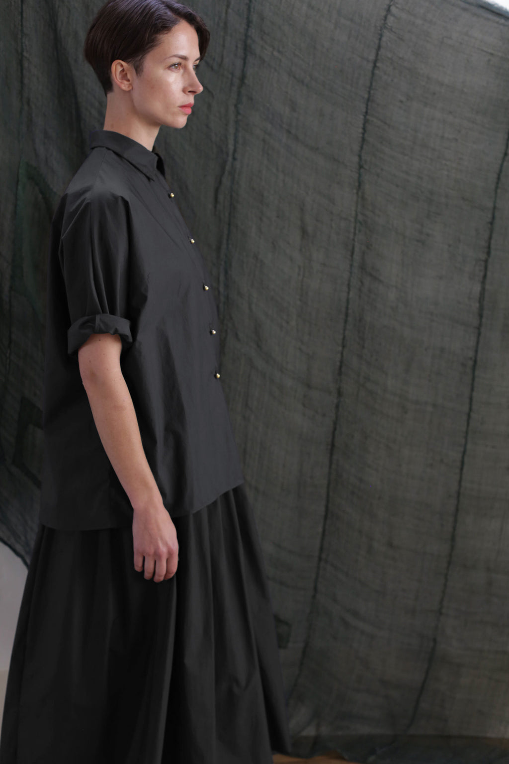 A woman with short brown hair stands in profile, wearing the Work Shirt | Typewriter Cotton Black. She is in front of a dark, semi-transparent fabric backdrop.